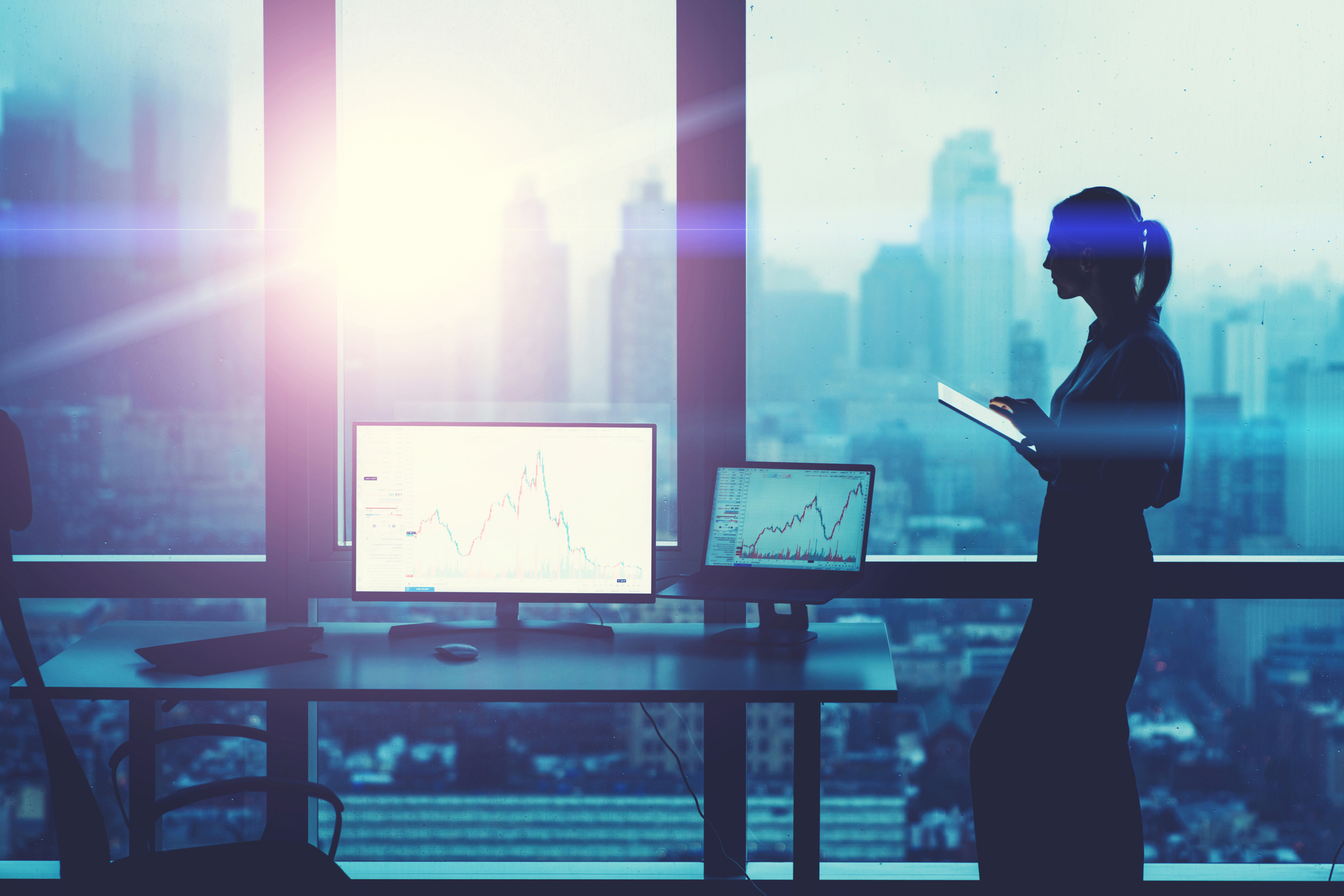 The silhouette of a businesswoman working on a tablet stands near a panoramic office window with a city view.
